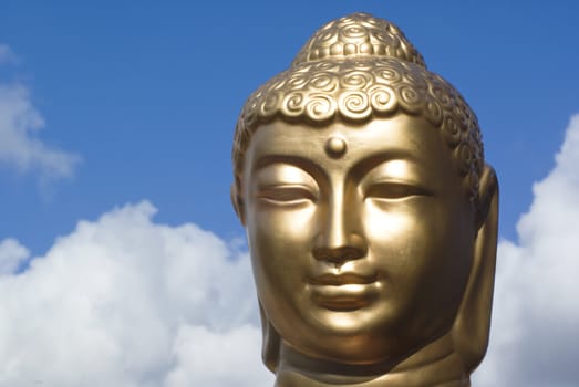 Head of a golden buddha against a blue cloudy sky.