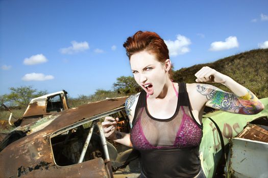 Angry tattooed Caucasian woman yelling with fists clenched in junkyard.