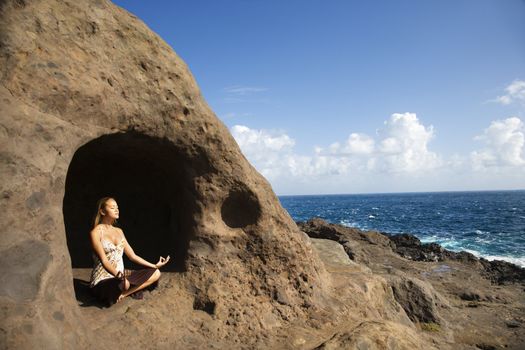 Young Asian woman mediatating in cave with coastline of Maui, Hawaii.