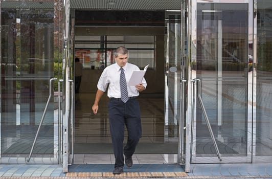 Businessman getting out of a corporate buidling while reading something in a notebook.