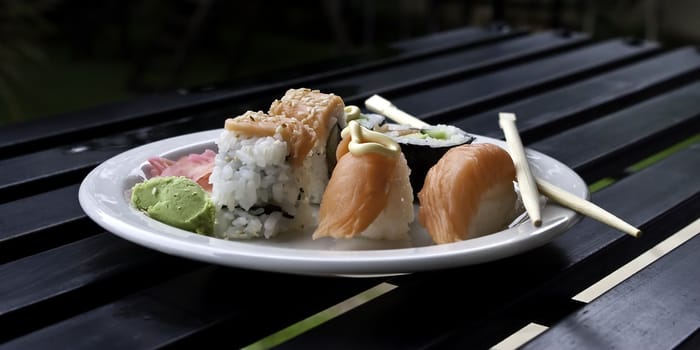 a plate of sushi on a white plate lying on a black wood table with eating utensils