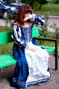 A girl in medieval blue dress sitting on a bench 
