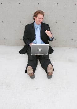 businessman sitting outside with his wireless laptop sitting on his lap while he's looking at his cell phone wearing a black suit and blue tie