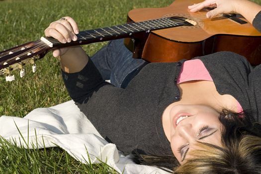 A young hispanic woman playing a guitar while laying on a blanket in the green grass.  Her hair is highlighted with blonde streaks.