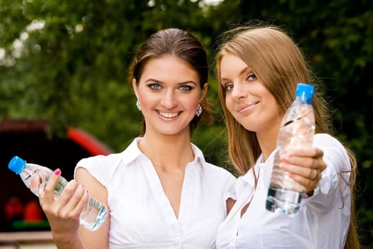 Two inseparable cheerful girlfriends holding bottle of water in summer park