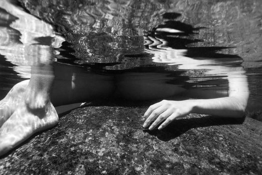 Caucasian young nude female arm and legs partially submerged underwater on rock.