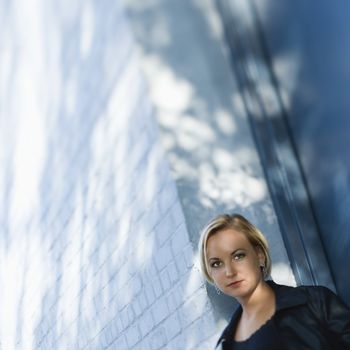 Caucasian mid-adult blonde woman standing against sun dappled building looking at viewer.