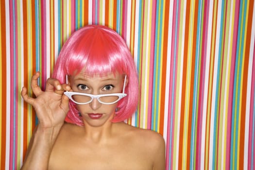 Portrait of attractive Caucasian young adult woman wearing pink wig against striped background peeking over glasses at viewer.