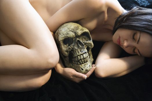 Nude Caucasian young adult woman lying down holding human skull.