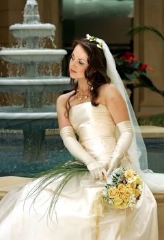 Beautiful bride in white dress holding bouquet and sat next to water fountain.