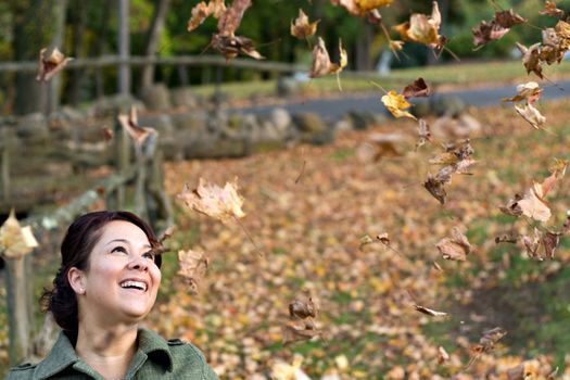 A pretty young woman laughing as the leaves fall all around her during the autumn season.