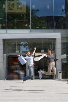 two young woman jumping in front of a commercial center with shopping bags