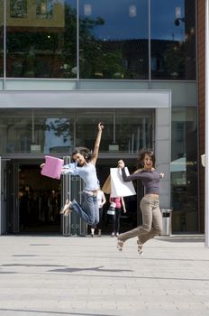 two young woman jumping in front of a commercial center with shopping bags