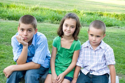 Three kids looking bored and overtired seated on a bench outdoors.