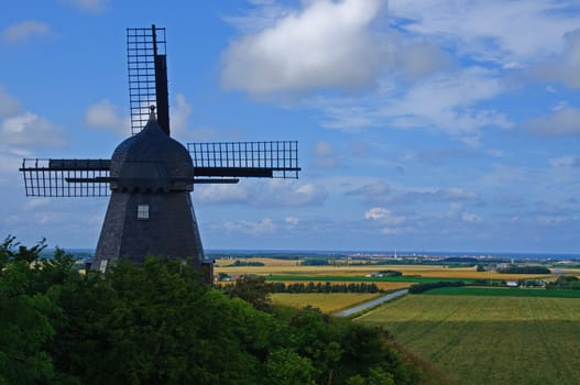 An old dark windmill on a  hill with a view over fields and to the ocean