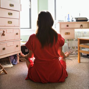 Back view of pretty Asian young woman in red robe sitting on floor meditating in lotus pose.