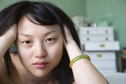 Portrait of pretty young Asian woman lying in bed with hands in hair making eye contact.