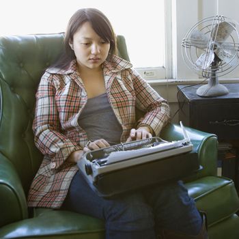 Pretty young Asian woman sitting in green chair typing on typewriter.