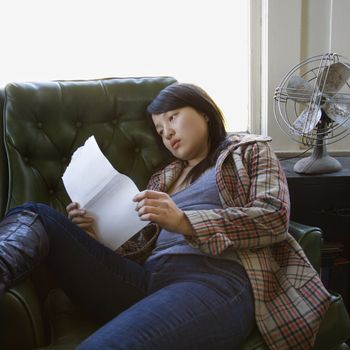 Pretty young Asian woman sitting in green chair reading paper.