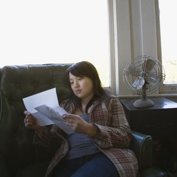 Pretty young Asian woman sitting in green chair reading letter.