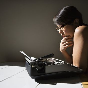 Nude young Asian woman sitting at kitchen table reading paper in typewriter.