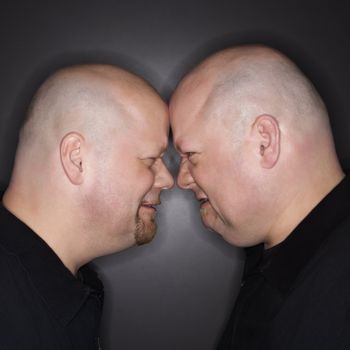 Caucasian bald mid adult identical twin men standing face to face with angry expression.