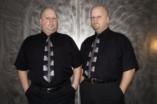 Caucasian bald mid adult identical twin standing together looking at viewer.