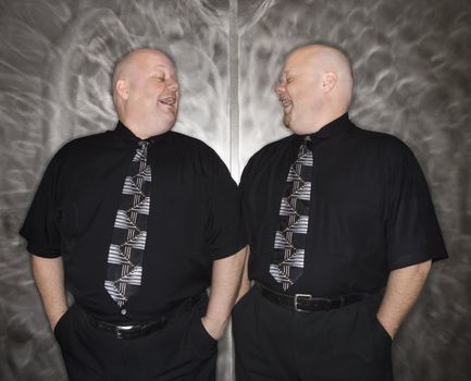 Caucasian bald mid adult identical twin men looking at each other laughing.