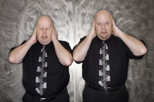 Caucasian bald mid adult identical twin men standing with hands covering ears making facial expression.