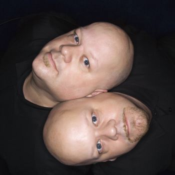 Aeriel view of Caucasian bald identical twin men sitting back to back and looking up at viewer.
