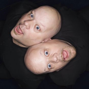 Aeriel view of Caucasian bald identical twin men sitting back to back making facial expressions.