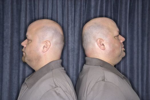 Profile of Caucasian bald identical twin mid adult men standing back to back.