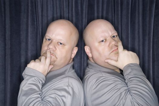 Caucasian bald identical twin men standing back to back and looking at viewer with hands to mouth.