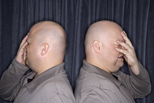 Profile of Caucasian bald identical twin men standing back to back and grimicing with hands to head.