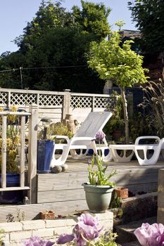 A small city garden setting with wooden constructed decking. Sun loungers rest on top of the decking surrounded by various ceramic plant containers, vegetation, flowers and plants.