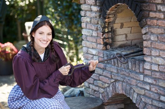Pretty farm girl dressed in traditional clothing baking bread in an outdoors oven