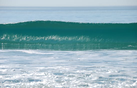 rising ocean wave shown on a sunny day with foam in the foreground