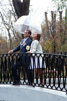 The groom and the bride stand on the bridge under an umbrella