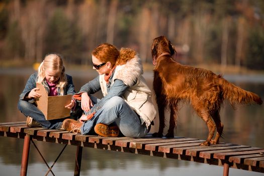 A girl reading a book with her mom at the lake in the forest, is a warm, sunny autumn day.