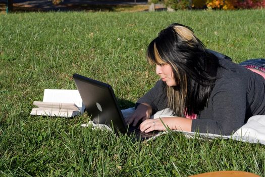 A young student using her laptop computer while laying in the grass on a nice day.