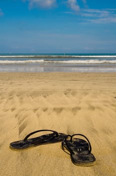 Sandals on sandy beach. In background wavy ocean.