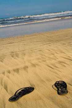 Sandals on sandy beach. In background wavy ocean.