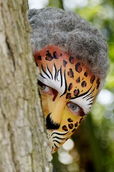 A man with his face painted like a leopard peeking around a tree.
