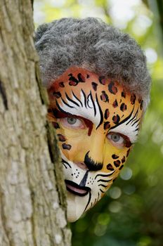 A man with his face painted like a leopard peeking around a tree.