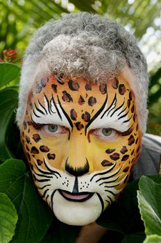 A man with his face painted like a leopard peeking out of large leaves.