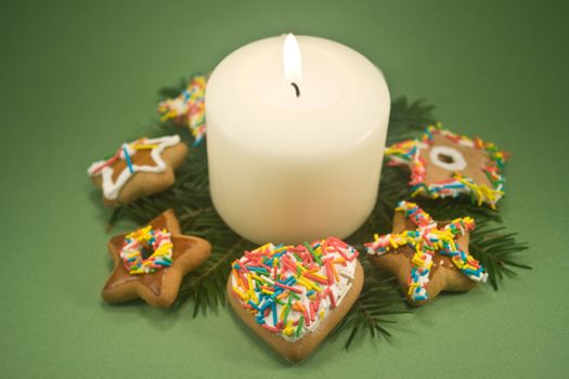 White pillar candle on fir branches surrounded by decorated gingerbread cookies on green background