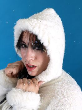 Beautiful brunette in a white hooded sweater, artificial snow falling down around her, against a blue background.