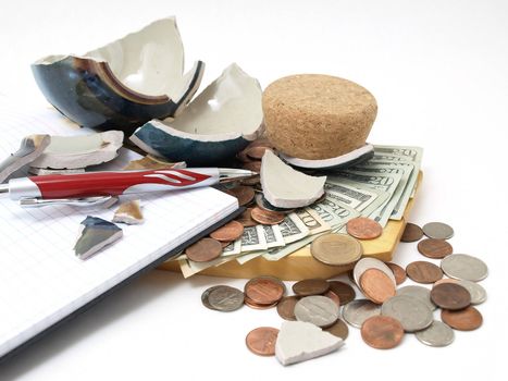 An open ledger and pen next to a broken ceramic bank on a pile of loose United States coins and currency.
