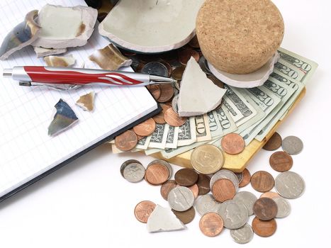 An open ledger and pen next to a broken ceramic bank on a pile of loose United States coins and currency.