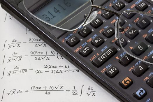 Scientific calculator with pi number on a display and reading glasses against math text book with integral tables. These are real devices, not props, showing some wear and dust. 